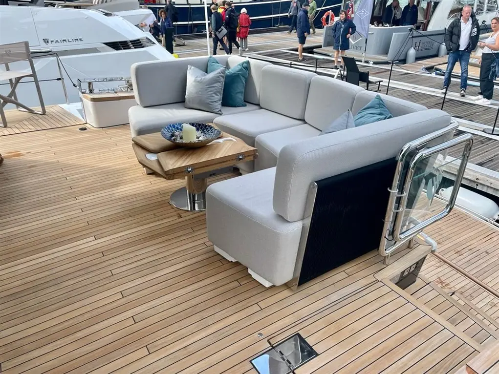Luxury yacht deck featuring a modern lounge area with a grey sofa and wooden table, surrounded by onlookers.
